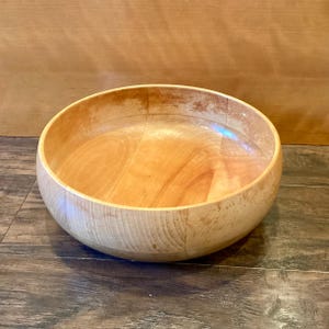 May include: A light-colored wooden bowl with a wide, shallow design. The bowl has a smooth, polished finish and a natural wood grain pattern. The bowl is suitable for serving salads or displaying decorative items.