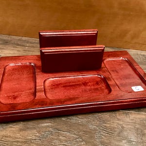 May include: A reddish-brown wooden desk tidy with several compartments. It has a rectangular base with three recessed sections and two raised, rectangular holders. The organiser is designed to hold various desk items.