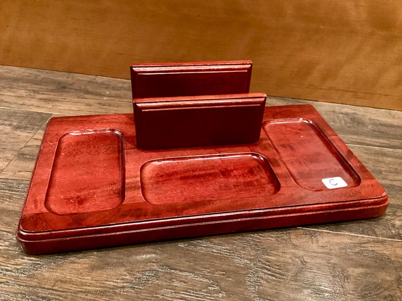 May include: A reddish-brown wooden desk tidy with several compartments. It has a rectangular base with three recessed sections and two raised, rectangular holders. The organiser is designed to hold various desk items.