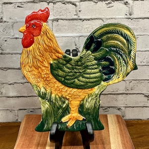 May include: A ceramic rooster-shaped decorative plate with a vibrant color scheme. The rooster features a red comb and wattle, a yellow beak, and a body with shades of yellow, green, and brown. The plate is supported by a black stand.
