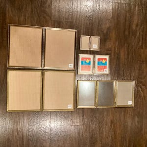 May include: A collection of gold-framed picture frames of various sizes, arranged on a wooden surface. Some frames have a textured backing, while others have a smooth surface. The frames are empty, ready for photos.