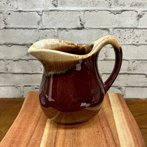 May include: A brown ceramic pitcher with a handle and a spout. The pitcher has a two-tone glaze, with a lighter tan color at the top and a darker brown color on the body. The pitcher is sitting on a wooden cutting board.