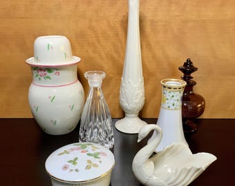 May include: A collection of seven decorative ceramic and glass items, including a swan figurine, a vase, a bottle, a jar, and a small box. The items are all white with floral designs and are arranged on a wooden surface.