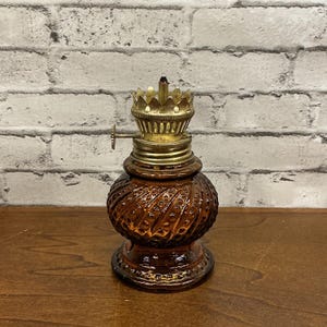 May include: An amber-colored glass oil lamp featuring a textured, swirled hobnail pattern on the base. The lamp includes a metallic brass-toned burner assembly with a crown-shaped gallery and a small adjustment wheel on the side for the wick.
