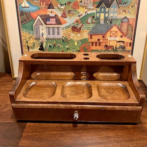 May include: A dark wooden desk tidy with several compartments and a small drawer. The organiser has a dark brown finish and features various shaped trays and slots for storage. A framed landscape painting is in the background.