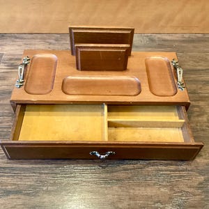 May include: Wooden desk tidy with a drawer and several compartments. The organiser is made of light brown wood and has a drawer with a decorative handle. It features slots for holding items and a section for letters or notes.