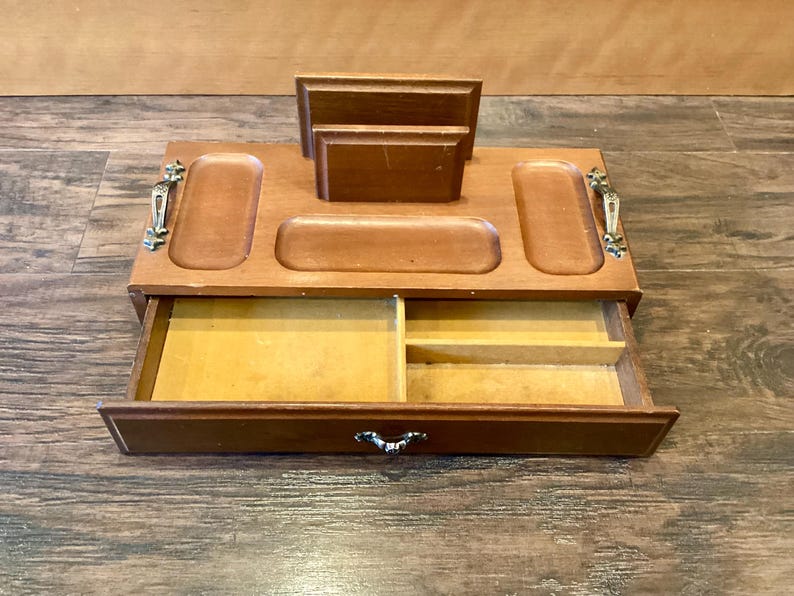 May include: Wooden desk tidy with a drawer and several compartments. The organiser is made of light brown wood and has a drawer with a decorative handle. It features slots for holding items and a section for letters or notes.