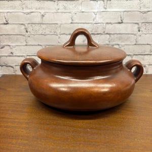 May include: A brown ceramic lidded casserole dish with two side handles and a loop handle on the lid. The dish has a glossy finish and sits on a wooden surface. The background is a white brick wall.
