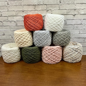 May include: A collection of ten yarn skeins in various colors, including orange, white, beige, gray, green, and pink. The yarn is arranged in a stacked formation against a white brick wall background. The skeins are on a brown wooden surface.