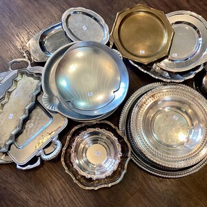 May include: A collection of vintage silver serving trays and dishes in various shapes and sizes. Includes oval, round, and octagonal trays, some with ornate edges and handles. The metal surfaces reflect light, creating a bright and elegant display.