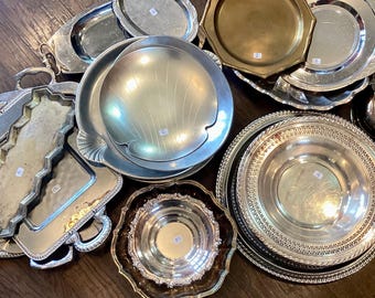 May include: A collection of vintage silver serving trays and dishes in various shapes and sizes. Includes oval, round, and octagonal trays, some with ornate edges and handles. The metal surfaces reflect light, creating a bright and elegant display.