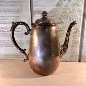 May include: A vintage silver-plated coffee pot with a curved handle and spout. The pot has a bulbous body and a lid with a decorative finial. The handle and spout feature ornate detailing. The pot is displayed on a light-colored wooden surface.