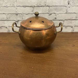 May include: A vintage copper pot with a lid and two handles. The pot has a round body and a lid with a small knob on top. The lid has three small holes. The pot is made of copper and has a warm, reddish-brown color.
