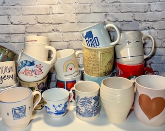 May include: A collection of various ceramic mugs, featuring diverse designs and colors. Some mugs have floral patterns, a rainbow, and the word "DAD". Others are plain or have simple designs. The mugs are stacked on a white surface.