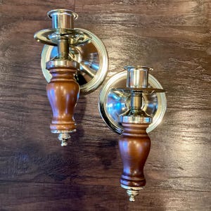 May include: Two wall-mounted candle holders. Each has a brass-colored circular backplate and a central column with a wooden, turned design. The candle cups are also brass-colored. The wood is a warm brown, and the metal has a polished finish.