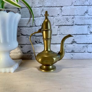 May include: A vintage brass pitcher with a long, curved spout and a handle. The pitcher has a bulbous body and a decorative top. The brass has a warm, golden hue, and the pitcher is likely used for pouring liquids.