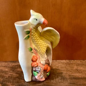 May include: A ceramic vase in the shape of a mythical bird with a yellow body, white head, and orange beak. The vase has a cream-colored base with a tree branch design, featuring orange fruit and green leaves. The vase is approximately 8 inches tall.