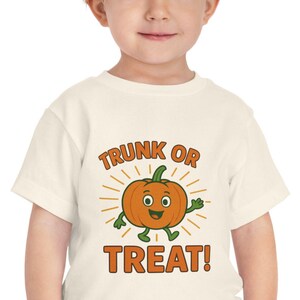 May include: A toddler wearing a cream-colored t-shirt with a cartoon pumpkin graphic. The pumpkin has a smiling face and the words "TRUNK OR TREAT!" in orange. The child is also wearing blue pants.