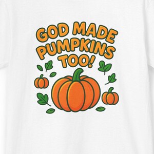 以下が含まれることがあります： オレンジ色のカボチャのグラフィック、緑の葉、オレンジと黄色のテキスト「GOD MADE PUMPKINS TOO！」が特徴の白いTシャツ。デザインには小さなカボチャが含まれています。