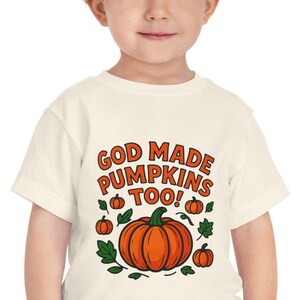 Pode incluir: Uma camiseta creme para crianças pequenas com o texto "GOD MADE PUMPKINS TOO!" em laranja. A camisa apresenta uma grande abóbora laranja com folhas verdes e abóboras menores. A criança está usando jeans azuis.