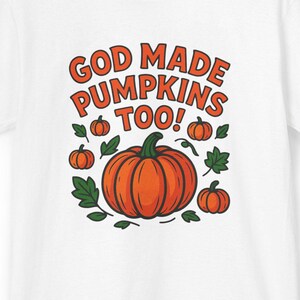 May include: White t-shirt with the text "GOD MADE PUMPKINS TOO!" in orange. The design features a large orange pumpkin surrounded by smaller pumpkins and green leaves, perfect for fall or Halloween.