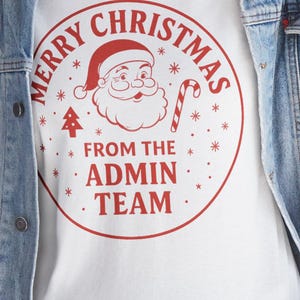 Op de afbeelding: Wit T-shirt met een rode cirkelvormige afbeelding met de tekst "Merry Christmas" en "From the Admin Team". De afbeelding bevat een kerstman, een zuurstok en een kerstboom. De persoon draagt een spijkerjasje.