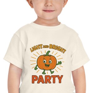 Może przedstawiać: Kremowy t-shirt dla malucha z uśmiechniętą dynią z kreskówki i napisem "LIGHT AND BRIGHT PARTY" w kolorze pomarańczowym i zielonym. Dynia otoczona jest żółtymi promieniami słońca. Dziecko ma na sobie niebieskie spodnie.