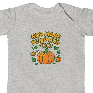 Op de afbeelding: Een lichtgrijze babybody met een afbeelding met de tekst "GOD MADE PUMPKINS TOO!" in geel en oranje. De afbeelding toont een grote oranje pompoen en twee kleinere pompoenen met groene bladeren.