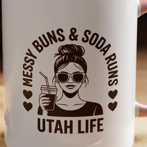 Puede incluir: Taza de cerámica blanca con interior y asa rosa. La taza presenta un gráfico de una mujer con gafas de sol y un moño desordenado, sosteniendo una bebida. El texto en la taza dice "Messy Buns & Soda Runs" y "Utah Life".