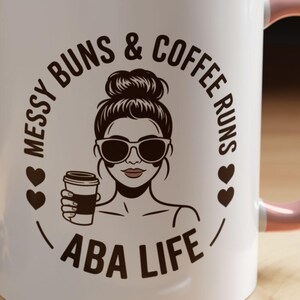 May include: White coffee mug with a pink interior and handle. The mug features a graphic of a woman with sunglasses and a bun, holding a coffee cup, surrounded by the text "Messy Buns & Coffee Runs ABA Life."