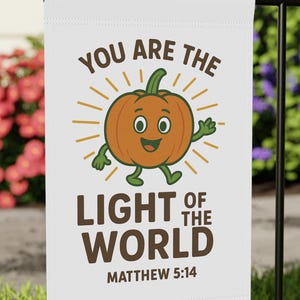 Könnte beinhalten: Eine weiße Gartenflagge mit einem Cartoon-Kürbis-Design und dem Text "YOU ARE THE LIGHT OF THE WORLD" und "MATTHEW 5:14". Der Kürbis ist orange mit einem grünen Stiel, Armen und Beinen. Die Flagge befindet sich auf einem schwarzen Metallständer.