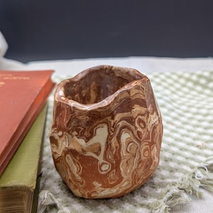 May include: A small, brown and white marbled ceramic planter with an irregular, textured surface. The planter is sitting on a green and white checkered fabric.