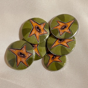 May include: A collection of round, green buttons with an orange star design. Each star features an eye in the center. The buttons are arranged on a light-colored surface.