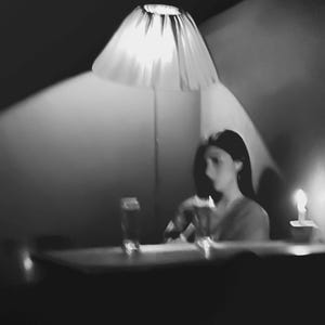 Könnte beinhalten: Eine Frau sitzt an einem Tisch in einem schwach beleuchteten Raum, der von einer Lampe mit einem weißen Lampenschirm beleuchtet wird. Sie schaut auf etwas in ihren Händen. Vor ihr steht ein Glas Wasser auf dem Tisch.
