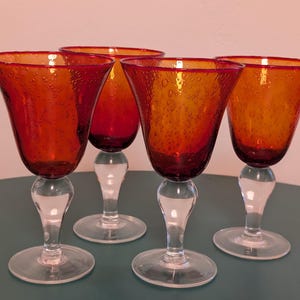 May include: A set of four wine glasses featuring deep red, bubble-textured glass bowls that transition into clear glass stems and circular bases. The bowls have a wide, flared rim and a distinct bulbous shape, showcasing a handcrafted appearance.