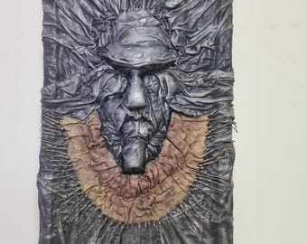 May include: A textured, abstract sculpture made of dark grey leather with a brown leather accent. The sculpture depicts a face with a closed mouth and eyes.