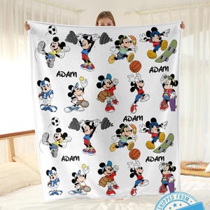 Puede incluir: Manta de forro polar blanca con un patr&oacute;n repetido de Mickey Mouse en varias poses deportivas. El nombre "ADAM" est&aacute; impreso en negro en la manta. Una persona sostiene la manta.