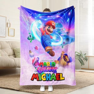 May include: A plush blanket featuring a vibrant graphic of Mario from "The Super Mario Galaxy" video game. The design includes Mario, various characters, and the name "MICHAEL" in colorful text against a purple and blue galaxy backdrop.