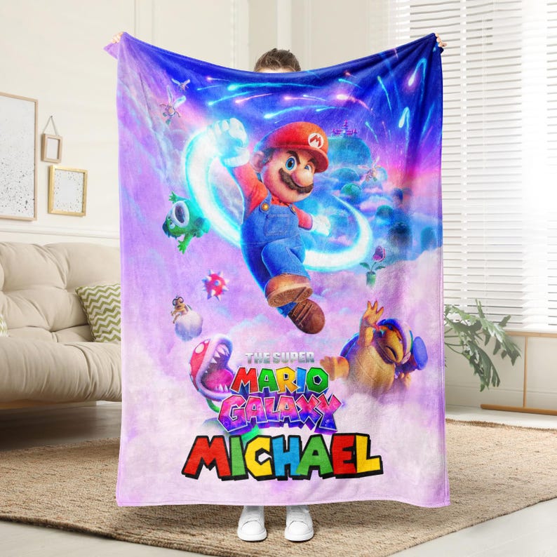 May include: A plush blanket featuring a vibrant graphic of Mario from "The Super Mario Galaxy" video game. The design includes Mario, various characters, and the name "MICHAEL" in colorful text against a purple and blue galaxy backdrop.