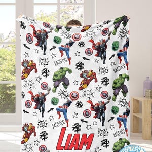 May include: White blanket featuring cartoon illustrations of superheroes like Thor, Captain America, Hulk, and Iron Man. The blanket has the name "LIAM" in red block letters and star accents. A fun, colorful design.