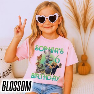 May include: A light pink t-shirt featuring a colorful graphic of characters from the movie Zootopia, with the text "SOPHIA'S 6 BIRTHDAY". The word "BLOSSOM" is at the bottom of the image.