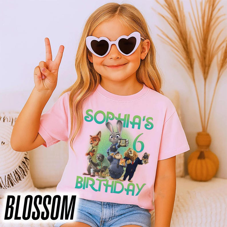 May include: A light pink t-shirt featuring a colorful graphic of characters from the movie Zootopia, with the text "SOPHIA'S 6 BIRTHDAY". The word "BLOSSOM" is at the bottom of the image.