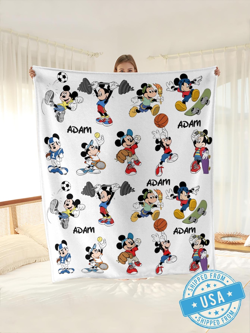 Puede incluir: Manta de forro polar blanca con un patr&oacute;n repetido de Mickey Mouse en varias poses deportivas. El nombre "ADAM" est&aacute; impreso en negro en la manta. Una persona sostiene la manta.