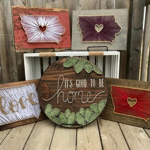 May include: Five wooden signs with string art designs. The signs feature a heart shape, the words "Love", "It's good to be home", and the outline of a state with a heart in the middle. The signs are all on a wooden surface.