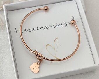 May include: Rose gold bangle bracelet with a knotted design and a small heart charm engraved with the letter "D". The bracelet is in a white box with the text "Herzensmensch" on the lid.