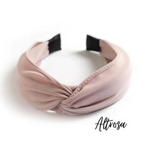 Könnte beinhalten: Ein hellrosa geknotetes Stirnband mit schwarzen Enden. Das Stirnband besteht aus einem weichen Stoff und hat ein verdrehtes Knotendesign. Das Wort "Altrosa" ist unten rechts in Kursivschrift geschrieben.
