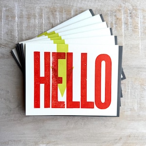 May include: A stack of white greeting cards with the word "HELLO" in bold red letters. A yellow arrow design is behind the "HELLO" text. The cards have gray envelopes and are arranged on a light wood surface.