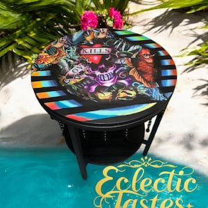 May include: A black side table with a round top featuring a colorful skull and tattoo-style artwork. The table has a lower shelf and decorative chain accents. The text "Eclectic Tastes by Design" is visible.