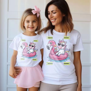 May include: Two white t-shirts featuring a unicorn wearing sunglasses and holding an ice cream cone, on a pink flamingo float. The shirts are worn by a mother and child. The image shows a matching set.