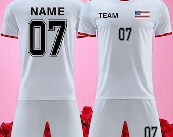 Puede incluir: Uniforme deportivo blanco con ribetes rojos, con el número 07 en negro. La camiseta tiene "TEAM" y una bandera estadounidense en la parte delantera, y "NAME" en la espalda. El pantalón corto es blanco.